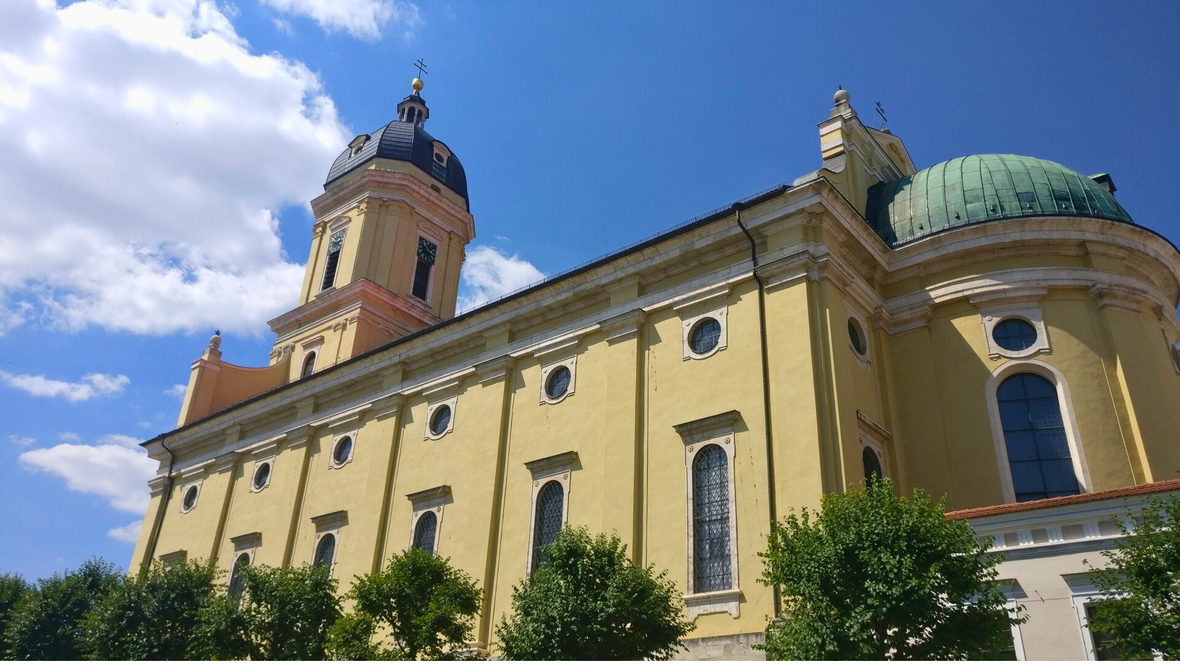 Außenansicht der Hofkirche Mariä Himmelfahrt in Neuburg an der Donau mit gelber Fassade, Turm und grüner Kuppel vor blauem Himmel.