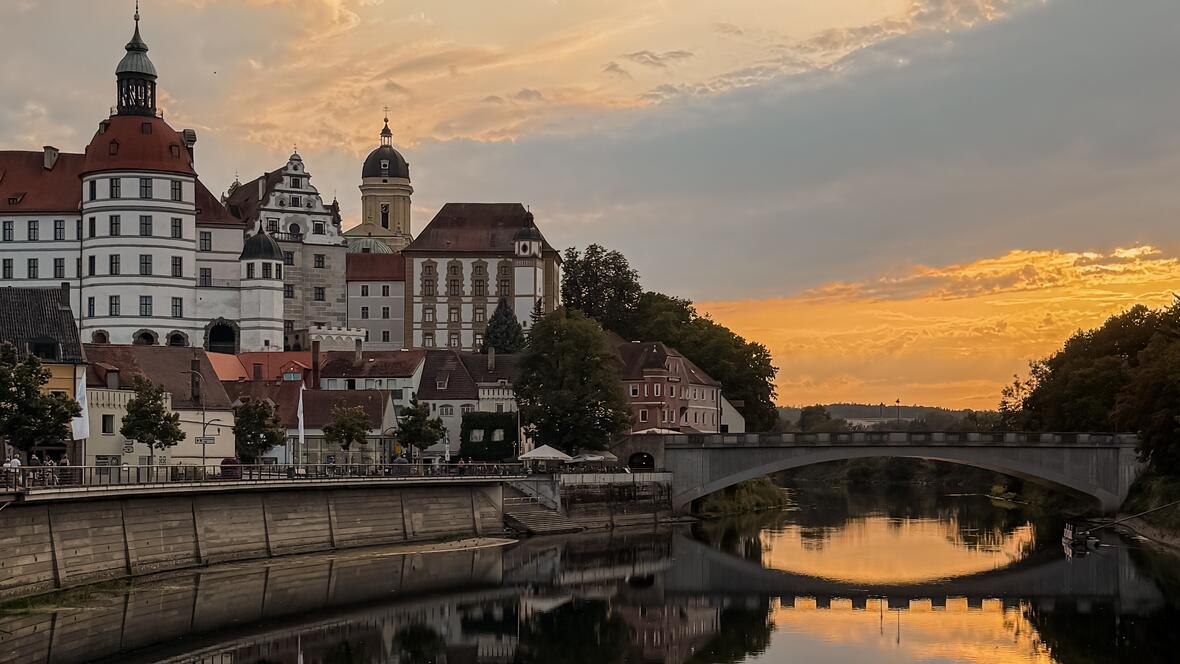 Das Neuburger Schloss spiegelt sich in der ruhigen Donau, während die untergehende Sonne den Himmel in goldenes Licht taucht.
