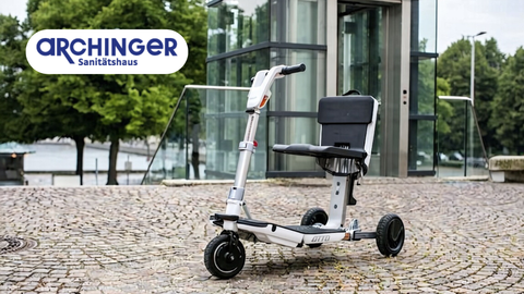 Elektromobil des Sanitätshauses Archinger in Neuburg auf einem gepflasterten Platz vor einem modernen verglasten Gebäude.